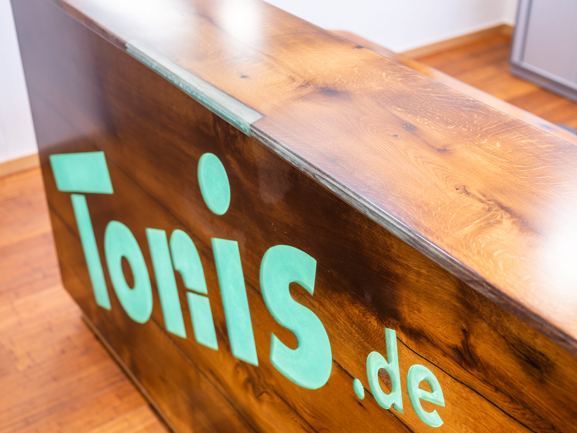 Holzempfangstheke mit dem grünen Logo von Tonis.de auf der Vorderseite.
